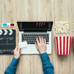 Photograph of a top-down view of a laptop with two hands on the keyboard. To the right of it are two popcorn bowls and boxes. To the left of it is a film set clapper.