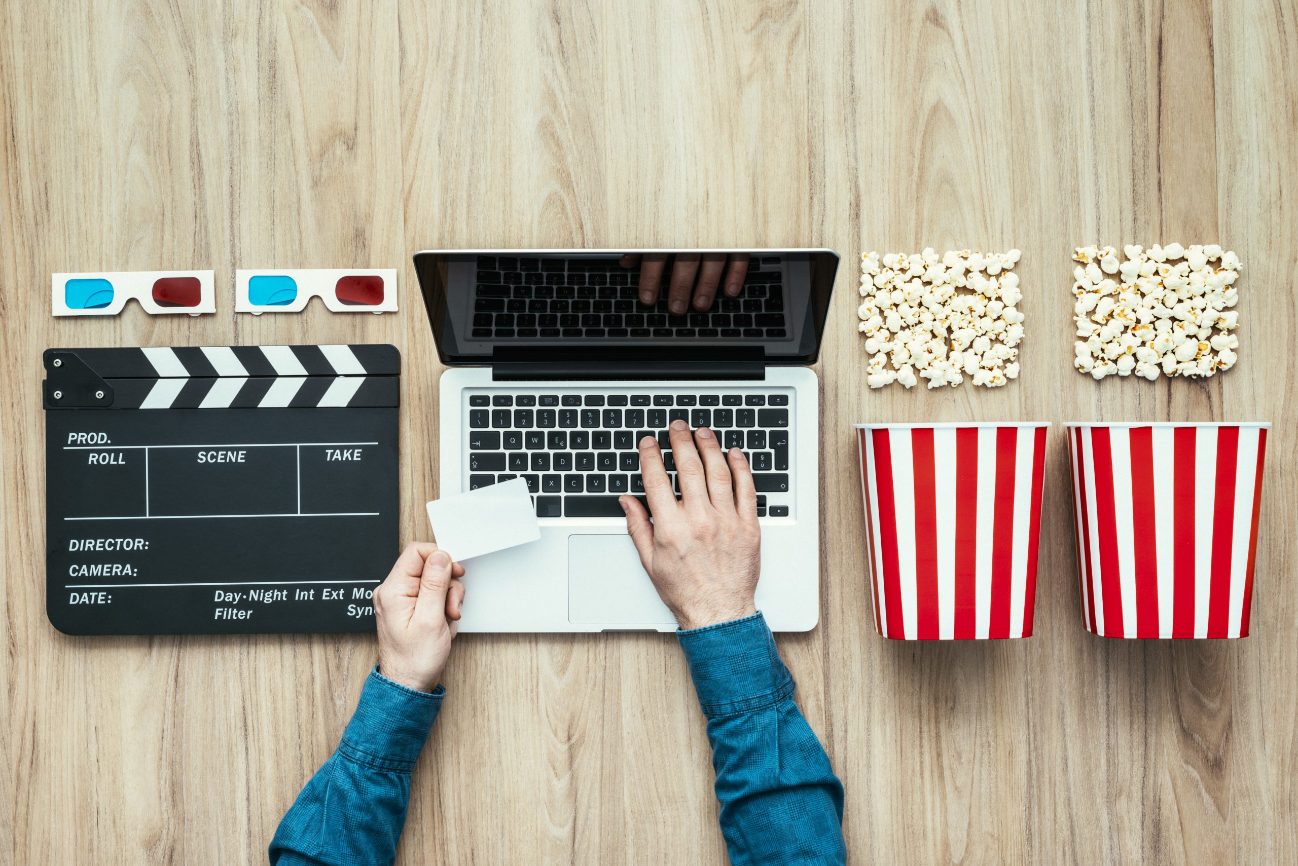 Photograph of a top-down view of a laptop with two hands on the keyboard. To the right of it are two popcorn bowls and boxes. To the left of it is a film set clapper.