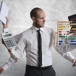 Photograph of a man looking distressed with six arms coming off of him, each holding a different item. The items include a planner book, a calculator, a magnifying glass, a laptop, an abacus, and a marker.