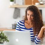 A beautiful woman with long brown, wavy hair sitting in front of her laptop wearing a blue and white striped shirt and holding out a credit card in her left hand.