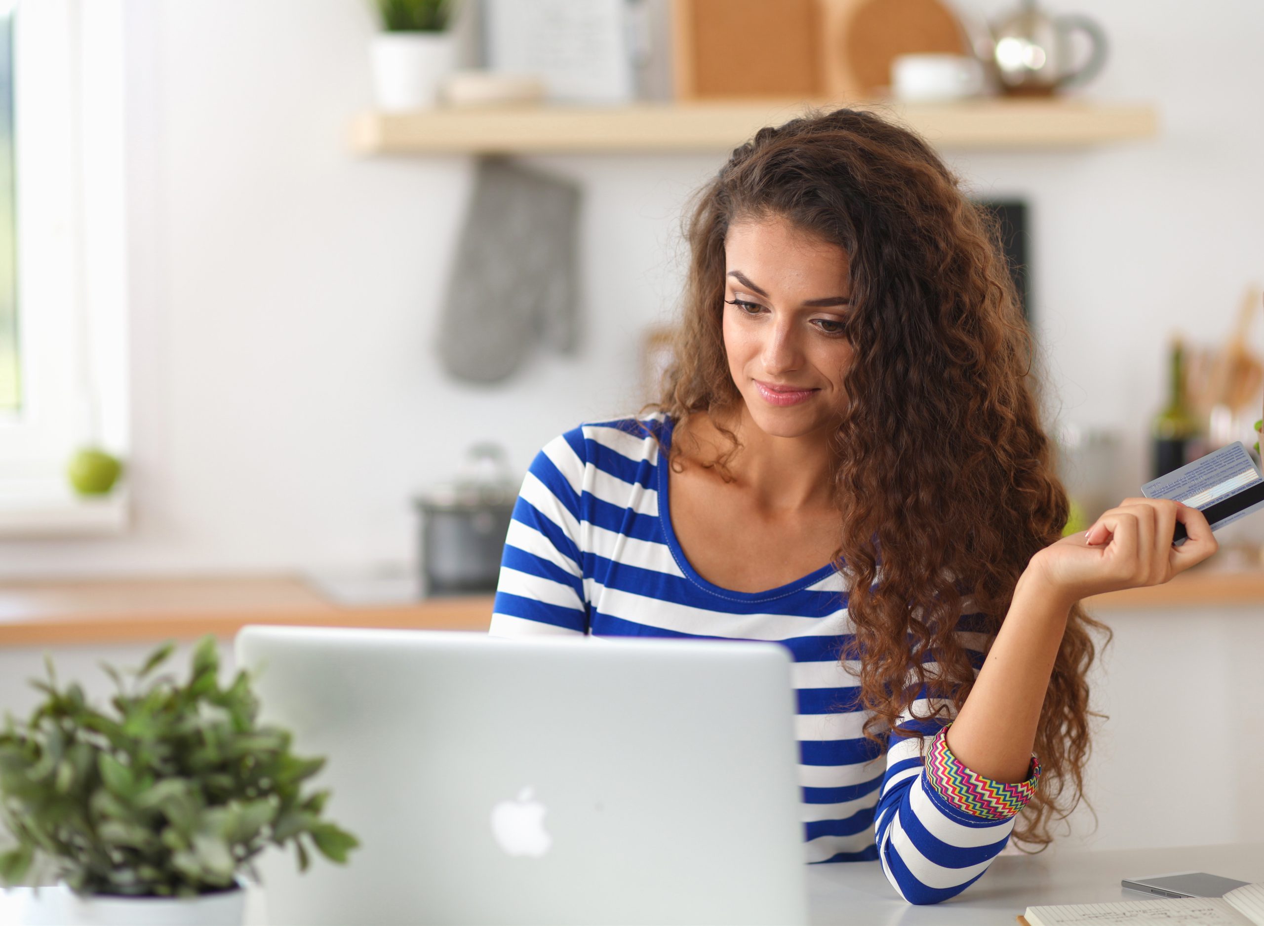 A beautiful woman with long brown, wavy hair sitting in front of her laptop wearing a blue and white striped shirt and holding out a credit card in her left hand.