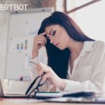 A beautiful woman with brown hair and her right hand to her forehead looking concerned. Her left hand is holding her glasses. She's looking down at her laptop. A chart with graphs is in the background.