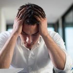 Image of a man with his hands on his head, looking down in a distressed manner with his eyes closed and a laptop sitting in front of him with the lid mostly closed.