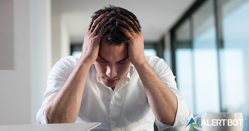 Image of a man with his hands on his head, looking down in a distressed manner with his eyes closed and a laptop sitting in front of him with the lid mostly closed.