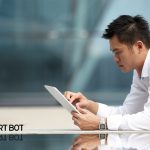 An asian man with spiky hair leans over a reflective table holding a tablet in his hand and is touching the screen with his right hand.