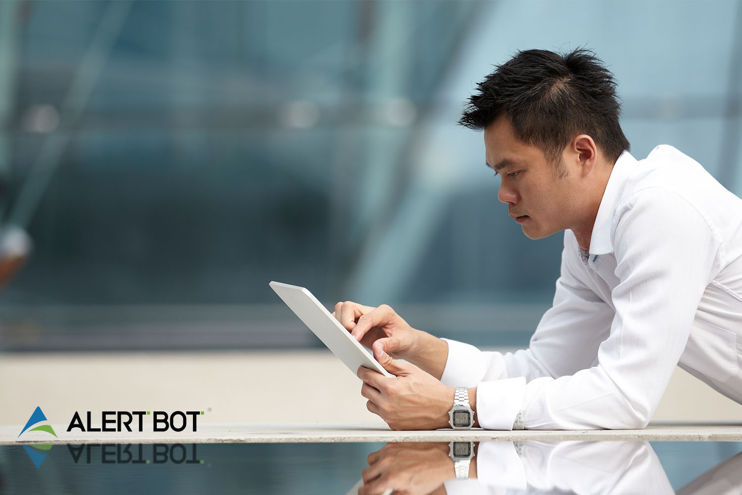 An asian man with spiky hair leans over a reflective table holding a tablet in his hand and is touching the screen with his right hand.