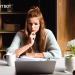 A pretty girl with brown hair up in a bun looks intently at a laptop with her hand to her chin in a pensive manner.