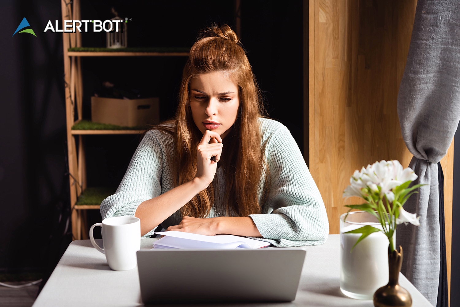 A pretty girl with brown hair up in a bun looks intently at a laptop with her hand to her chin in a pensive manner.