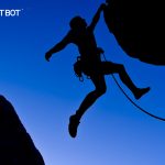 A mountain climber is silhouetted on a deep blue sky background as he hangs off a cliff by one hand, and a tether hangs off his belt.