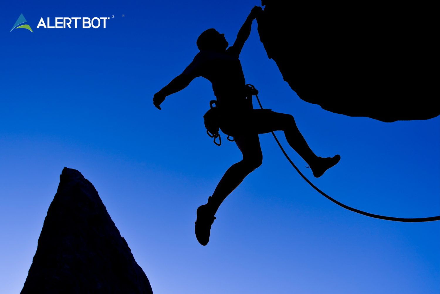 A mountain climber is silhouetted on a deep blue sky background as he hangs off a cliff by one hand, and a tether hangs off his belt.