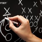 A hand holding chalk writes football plays on a blackboard, using x's, arrows and o's.