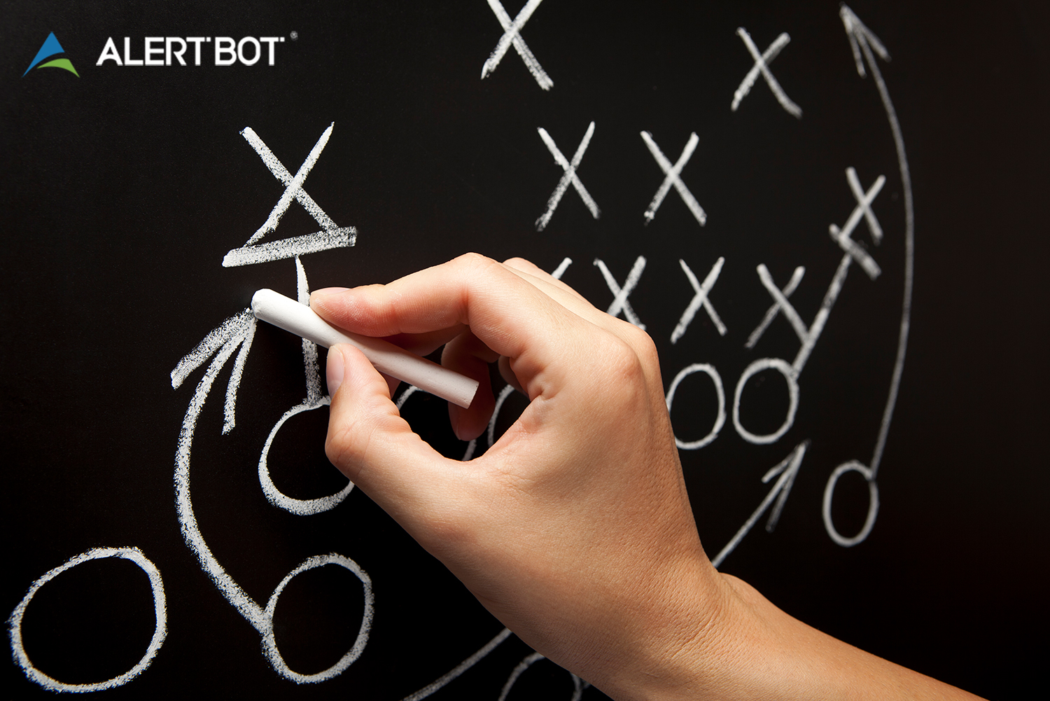A hand holding chalk writes football plays on a blackboard, using x's, arrows and o's.
