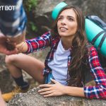 AlertBot blog titled "What is Proactive ScriptAssist and Why is it a Game-Changer?" with photo of a brown-haired woman in a white t-shirt and plaid button down shirt hiking and reaching up to grab the hands of someone helping to pull her up.