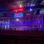 The BattleBots caged arena view from the stands. A monitor with the BattleBots logo hangs in front of the cage from the ceiling.