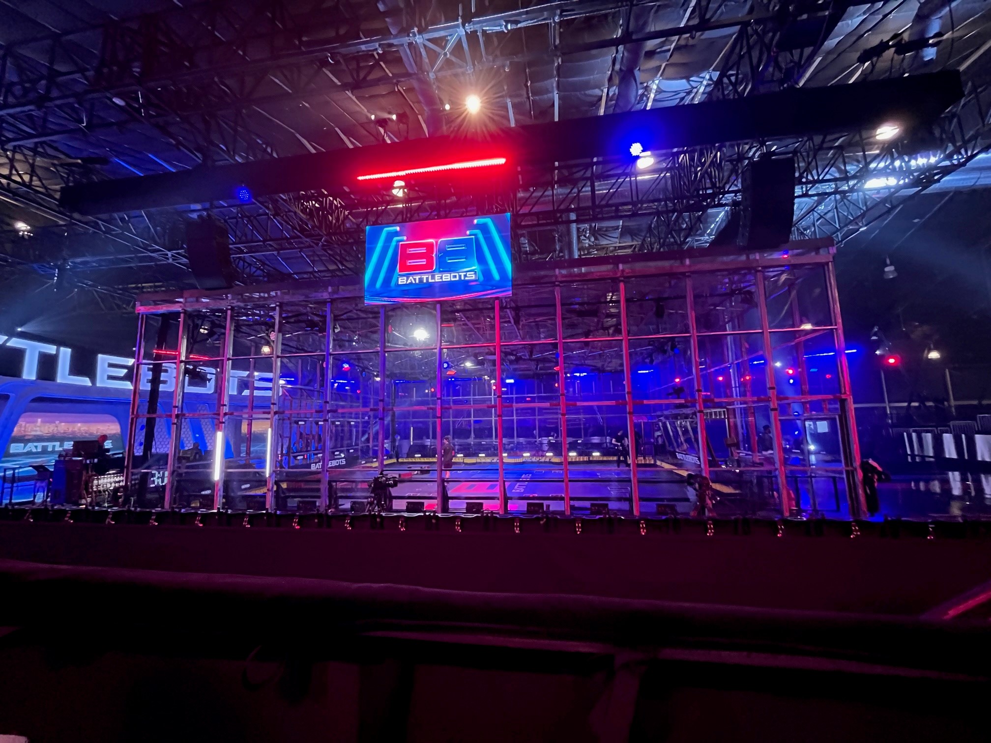The BattleBots caged arena view from the stands. A monitor with the BattleBots logo hangs in front of the cage from the ceiling.