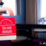 AlertBot blog titled "Why Your Website Monitoring Solution Needs a Do-Not-Disturb Feature" with a photo of a hotel room out of focus in the background and a "Do Not Disturb" sign on the doorknob in the foreground. AlertBot logo is placed in the lower right.