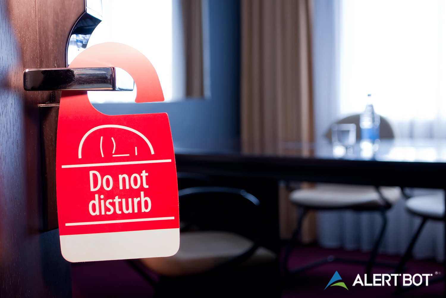 AlertBot blog titled "Why Your Website Monitoring Solution Needs a Do-Not-Disturb Feature" with a photo of a hotel room out of focus in the background and a "Do Not Disturb" sign on the doorknob in the foreground. AlertBot logo is placed in the lower right.