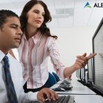 Woman with brown hair pointing at a computer monitor, while man with dark hair sits at a computer keyboard with hand resting on the keys.